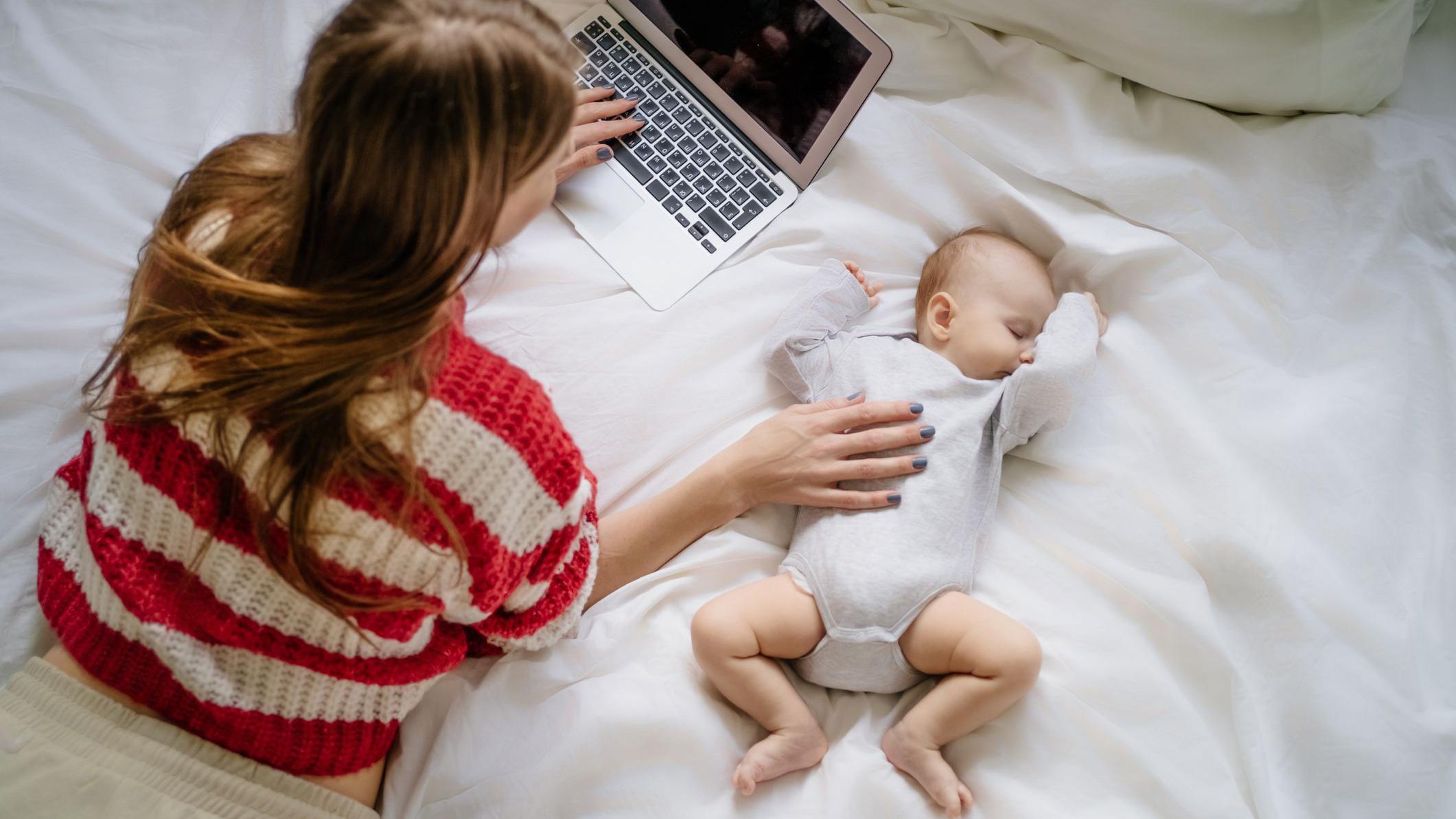 Foto de uma mulher deitada em uma cama de barriga para baixo, em frente a um computador e com uma mão na barriga de um bebê ao lado dela. 