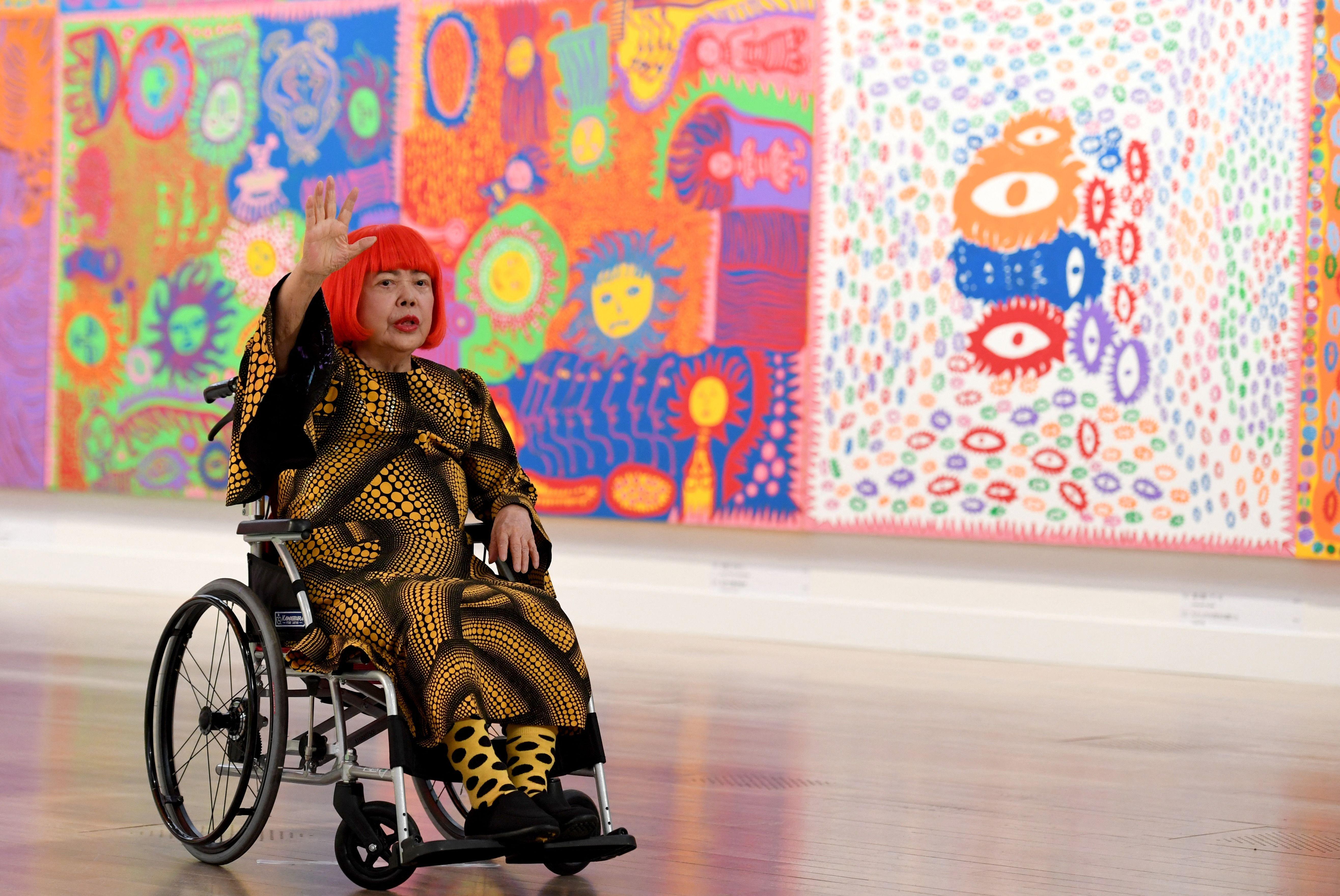 Kusama en silla de ruedas, con el pelo rojo y ropa de lunares negros y amarillos, frente a grandes pinturas multicolores.