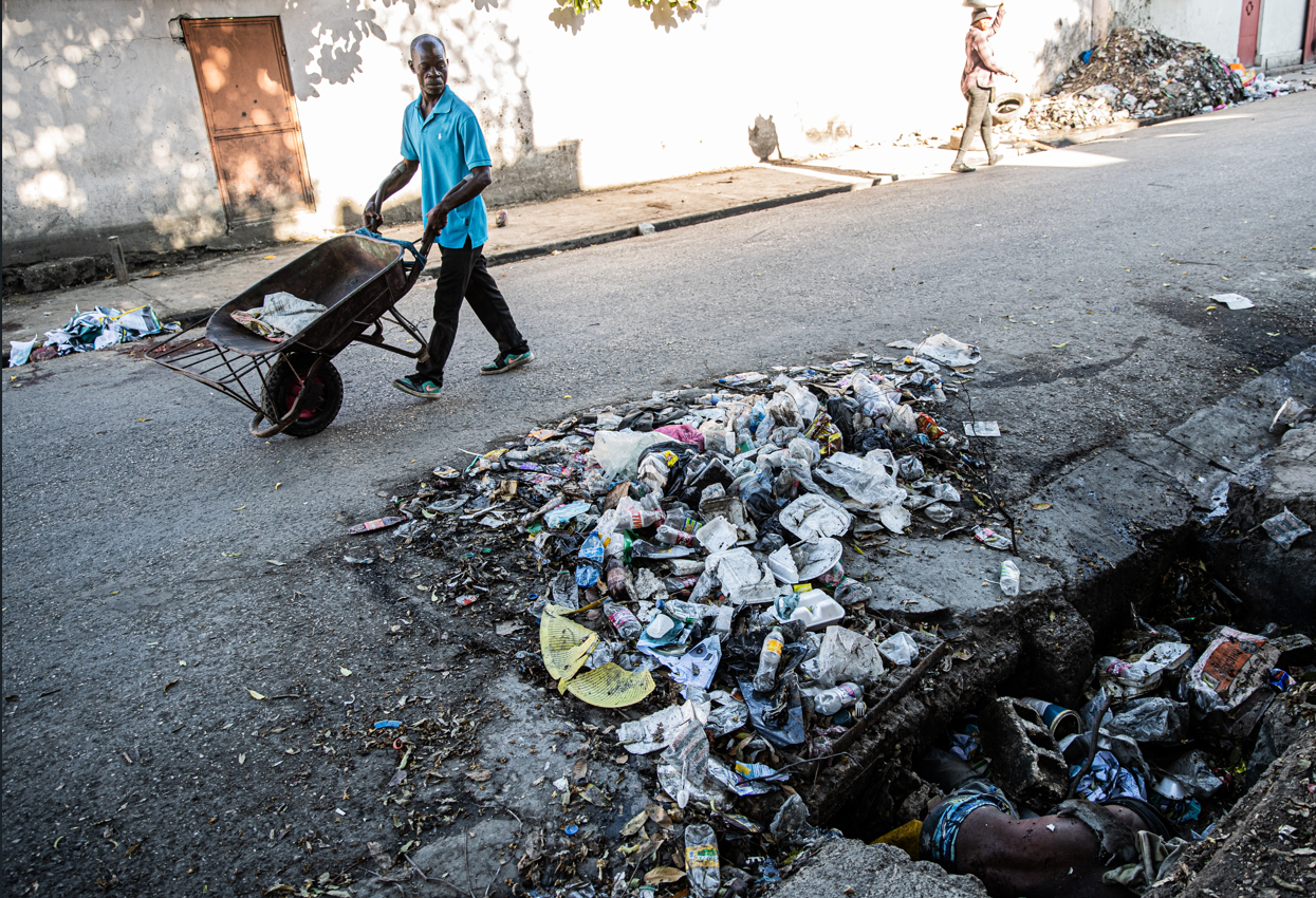 Un hombre con camisa azul pasa empujando una carretilla junto a un montón de basura y una zanja en la que se ve el torso desnudo de una persona muerta en Puerto Príncipe, Haití, el 19 de octubre de 2025.