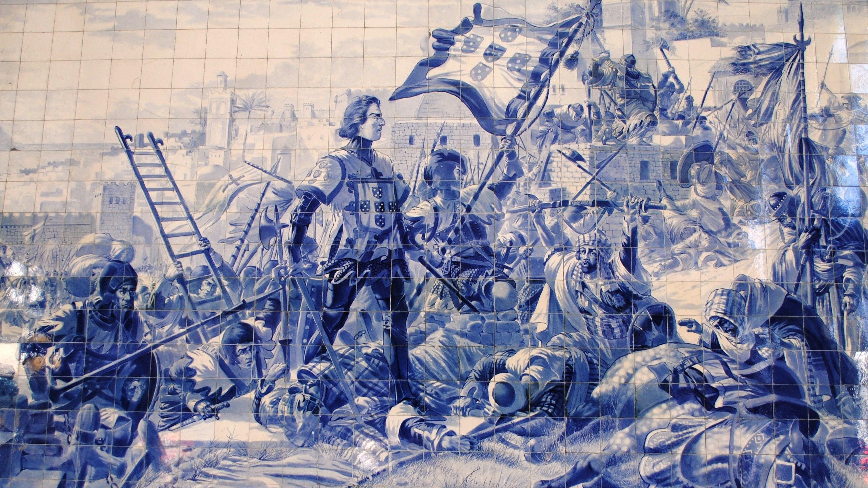 Painel em azulejos azuis e brancos retrata uma batalha medieval diante de uma cidade fortificada. Ao centro, um cavaleiro com a bandeira de Portugal lidera o combate, cercado por soldados armados, escadas de cerco, arqueiros e guerreiros ca&iacute;dos no campo de batalha.
