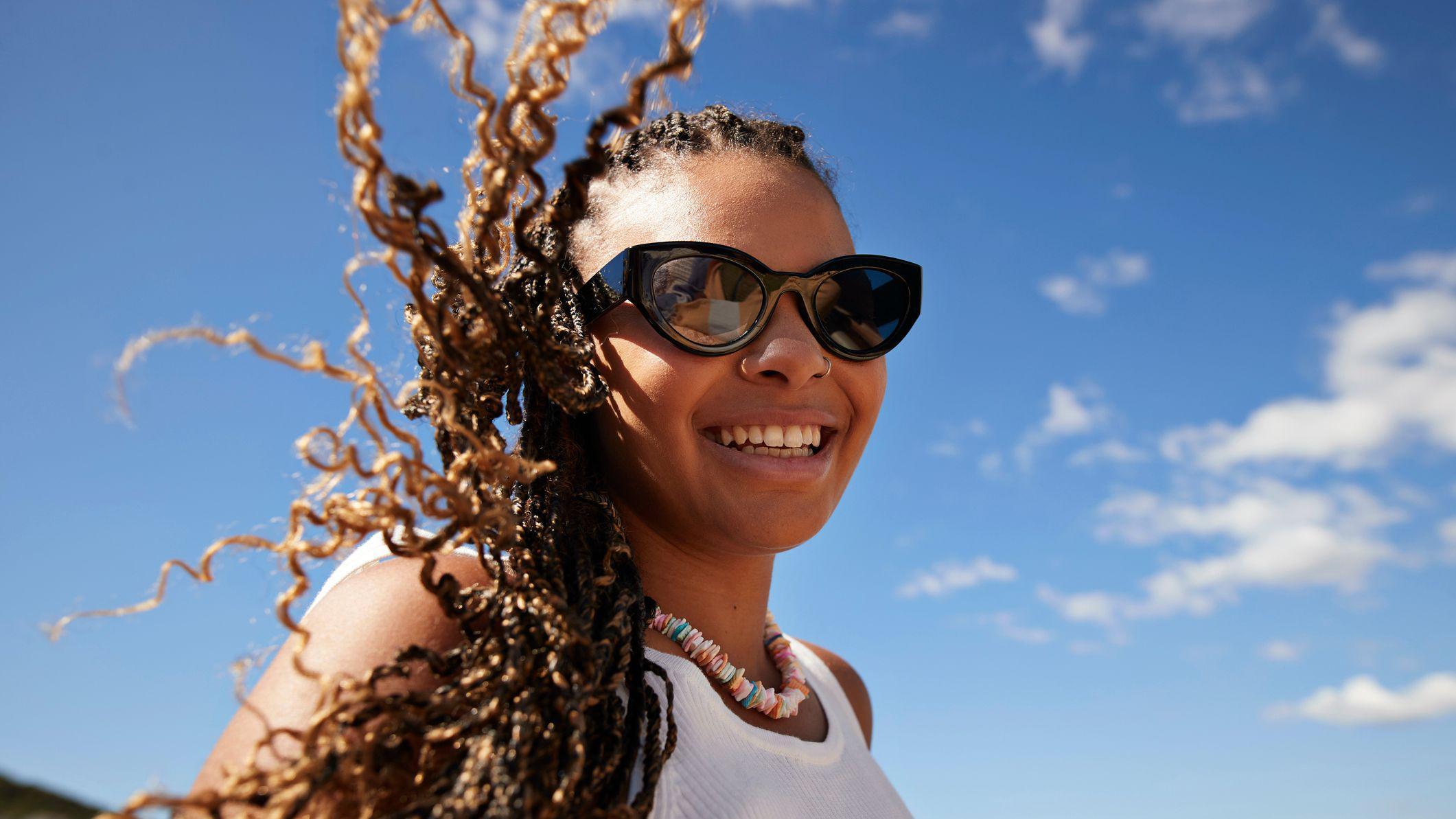 Mulher jovem de &oacute;culos escuros e cabelos esvoa&ccedil;antes em ambiente externo, em dia de sol