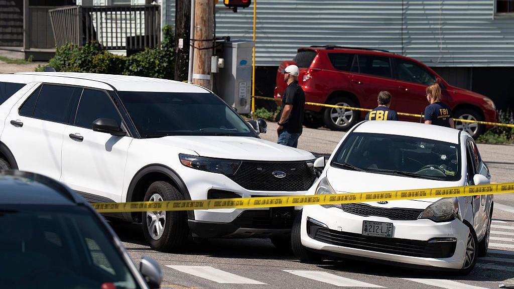 Dos coches blancos que colisionaron están detrás de cinta policial amarilla. El parabrisas de uno de los coches tiene agujeros de bala.