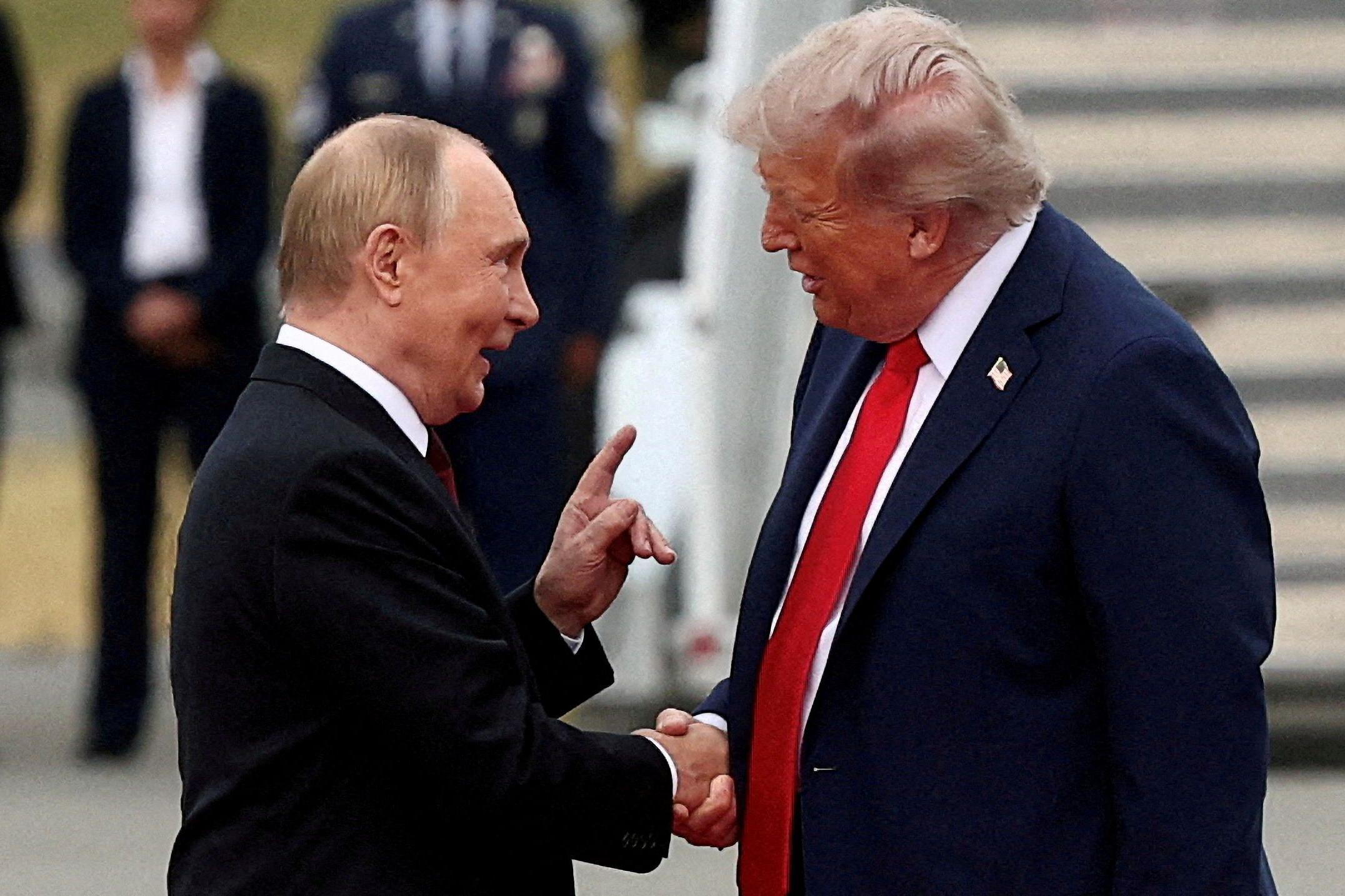 Putin y Trump se dan la mano frente a la escalerilla de un avión. Putin levanta la mano izquierda con el dedo índice apuntando hacia arriba y habla con Trump con la boca abierta. Hay gente al fondo; uno de ellos lleva condecoraciones militares. Trump viste una chaqueta oscura y corbata roja. Putin lleva una chaqueta negra. Ambos visten camisa blanca.
