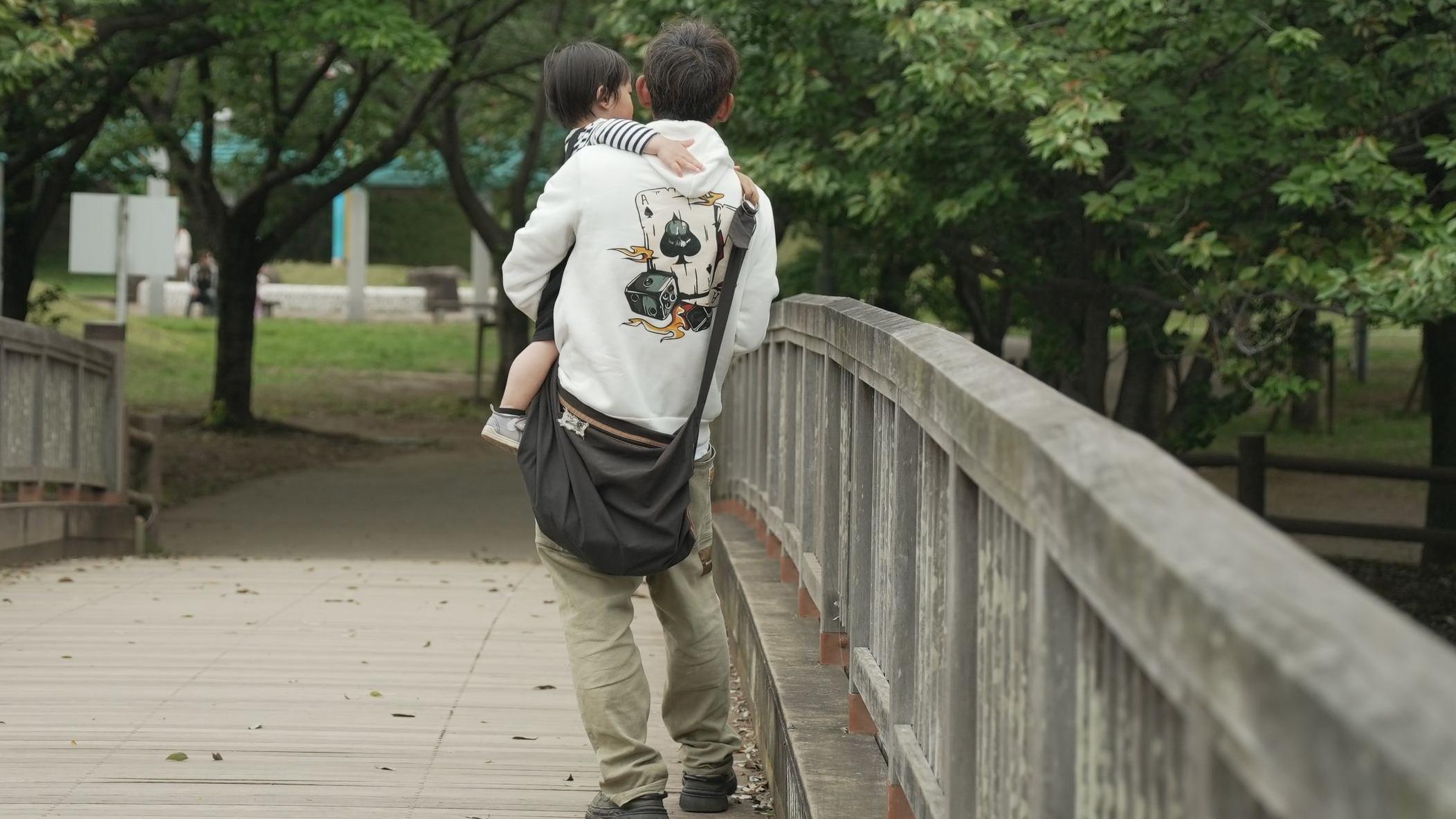 一名身穿連帽上衣的男子抱著嬰兒走過公園裡的一座橋。