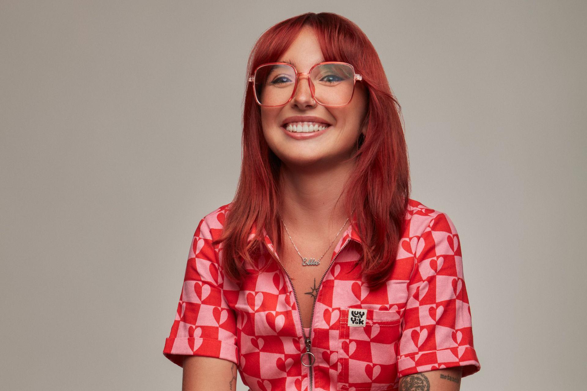 Ellie Middleton, de cabelos longos e ruivos e óculos, sorri contra um fundo cinza simples