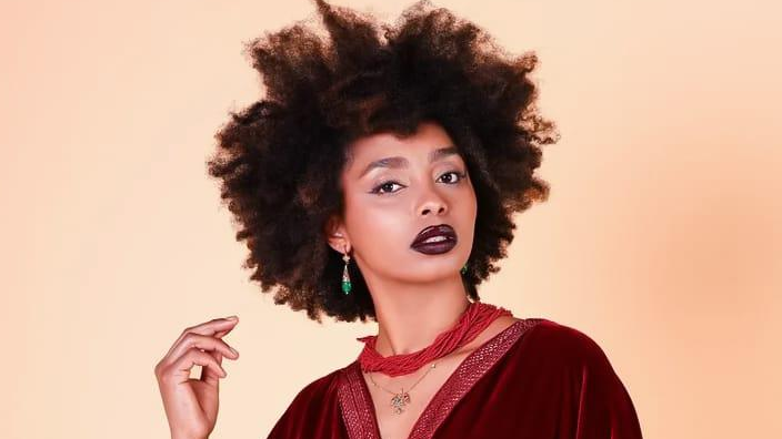 Foto de busto da modelo e criadora de conte&uacute;do Houda Fonone, vestindo uma blusa vermelha escura, um colar de contas vermelhas e brincos verdes.