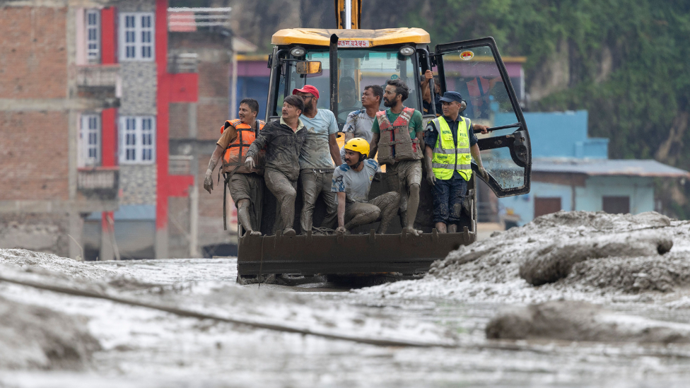 Un grupo de rescatistas y trabajadores de recuperación, cubiertos de barro, viajan en la pala delantera de una excavadora amarilla entre lodo espeso y escombros. Varios de ellos llevan chalecos reflectantes de alta visibilidad, y uno lleva un casco de seguridad amarillo.