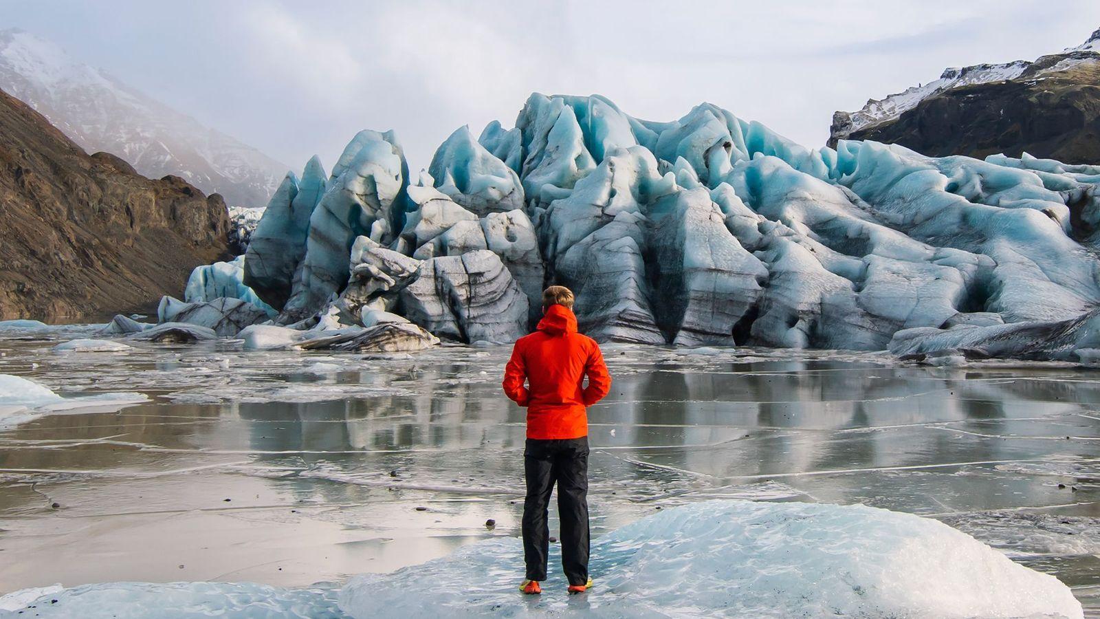Una persona con un abrigo rojo y pantalones negros, está parada sobre una superficie congelada observando un panorama de glaciares y montañas