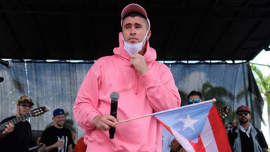 Bad Bunny con sudadera rosa y un micrófono sostiene una bandera de Puerto Rico con el triángulo celeste.