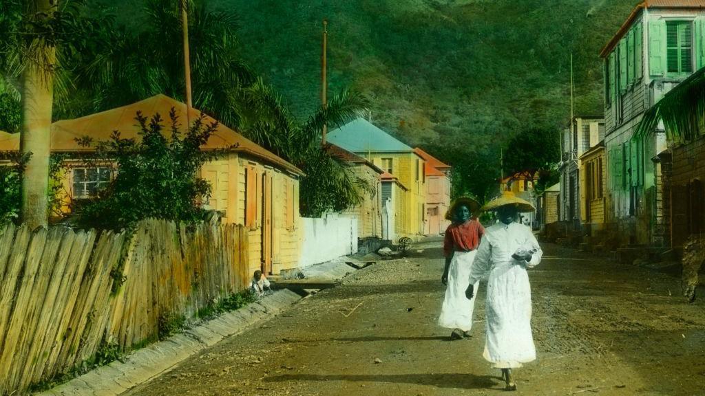 Duas pessoas usando saias e chap&eacute;us caminham por uma rua em St. Thomas, nas Ilhas Virgens Americanas, em uma foto de cerca de 1890