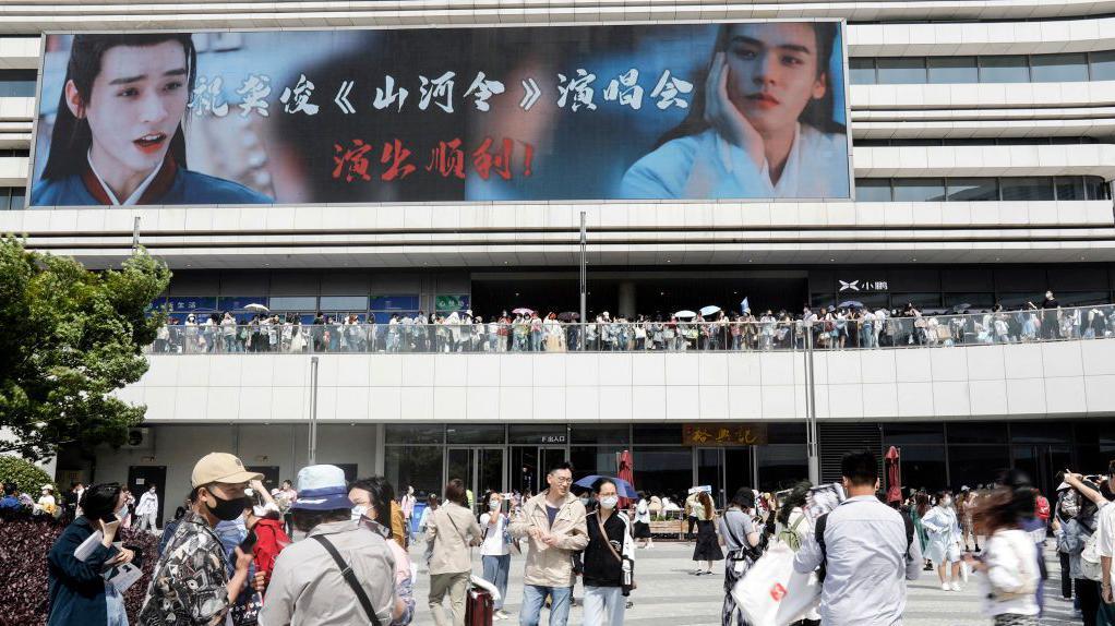 Fila de pessoas em frente a um est&aacute;dio com um painel que tem a propagada do drama 