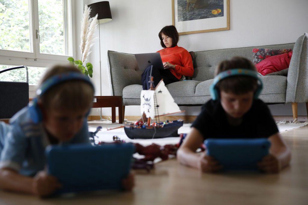 Duas crianças deitadas no chão com iPads e uma mãe sentada no sofá usando o computador