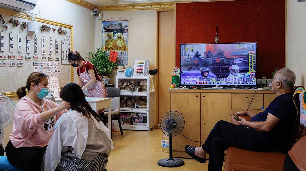 A television shows a news broadcast reporting on Chinese military drills around Taiwan, as staff and customers are seen inside a traditional facial threading shop in Hsinchu on December 29, 2025. China launched "major" military exercises around Taiwan on December 29, in what it called a "stern warning" involving live-fire drills in waters and airspace near the island. Taiwan said Monday it had detected four Chinese coastguard ships near the island's waters