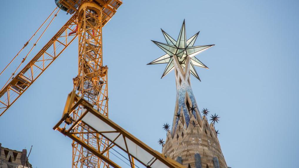 Una grúa de construcción amarilla se alza junto a la parte superior de una torre, que tiene una estrella de 12 puntas.
