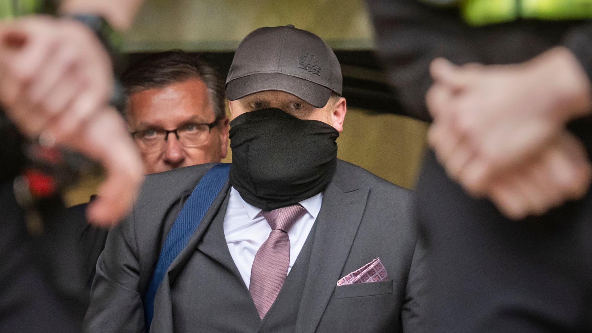 Bivši direktor pogrebnog preduzeća Robert Buš napušta zgradu Kraunskog suda u Halu, noseći sivo odelo sa ljubičastom kravatom i maramicom za džep.
Nosi tamnosivi kačket i crni šal, koji mu je omotan oko većeg dela lica ispod očiju.