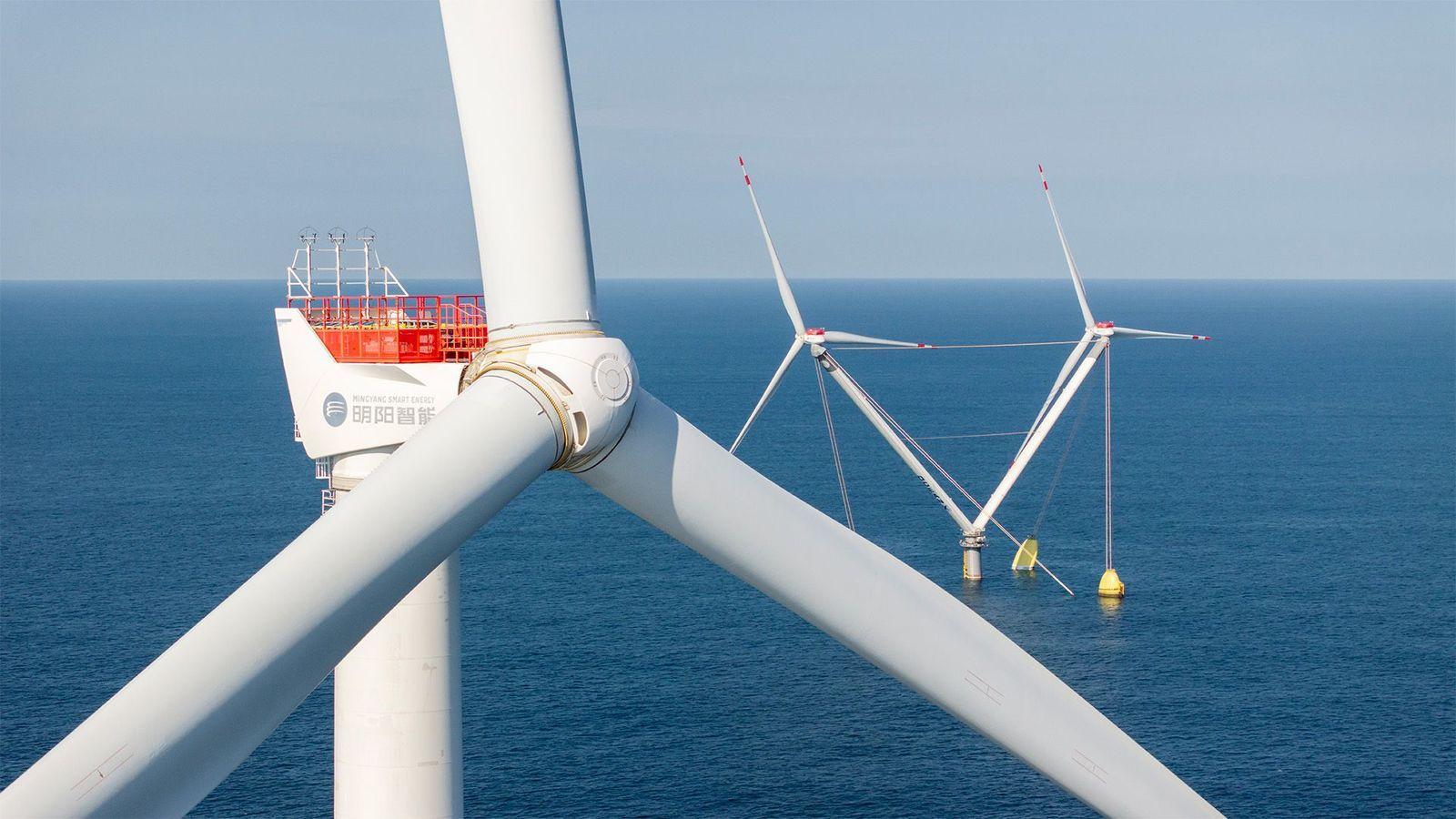 Plano cerrado del eje de las aspas de una turbina eólica marina blanca en el mar azul, con una torre de dos turbinas en el fondo