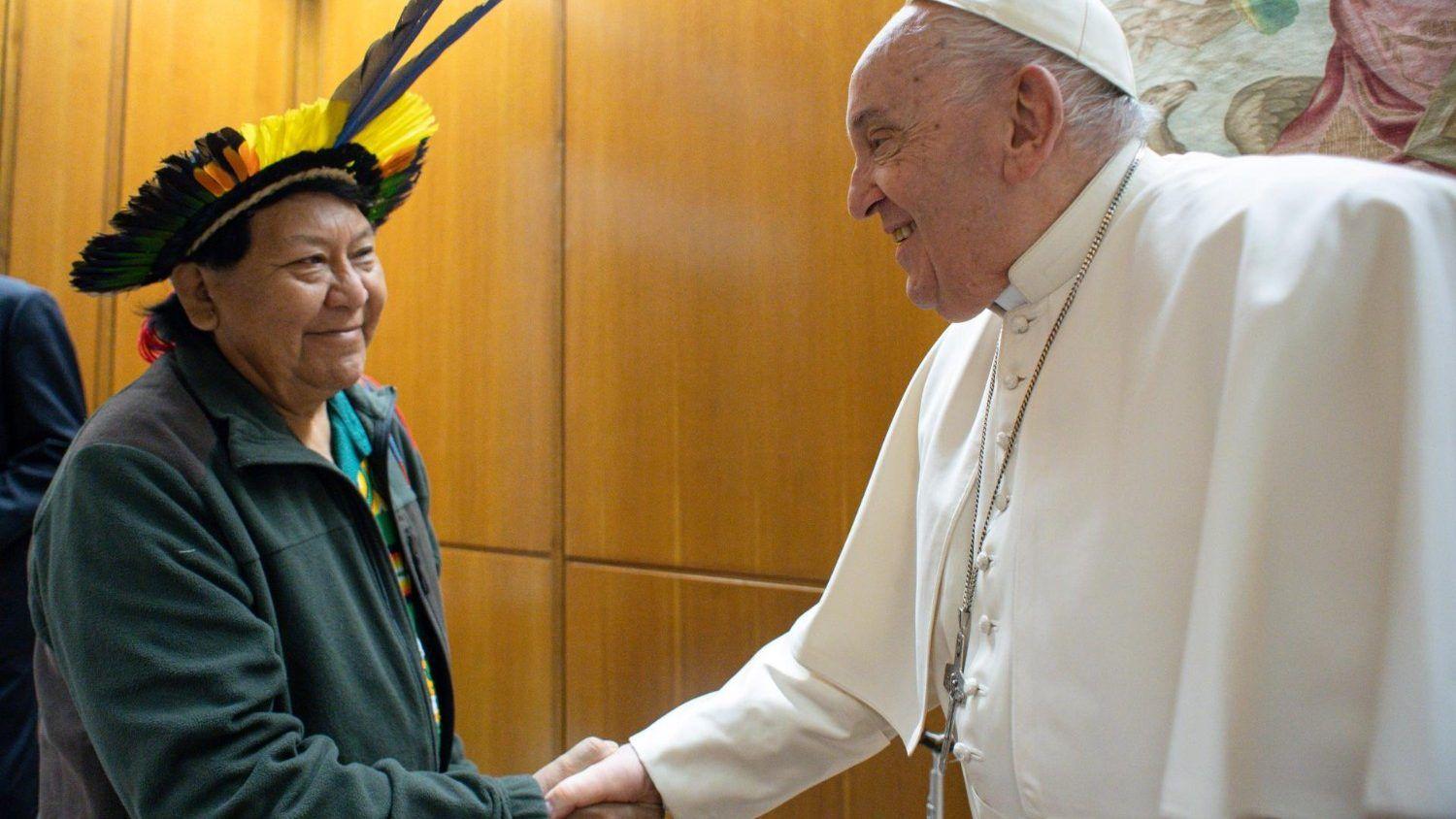 Francisco, com trajes brancos, cumprimenta homem que veste cocar com penas de aves.