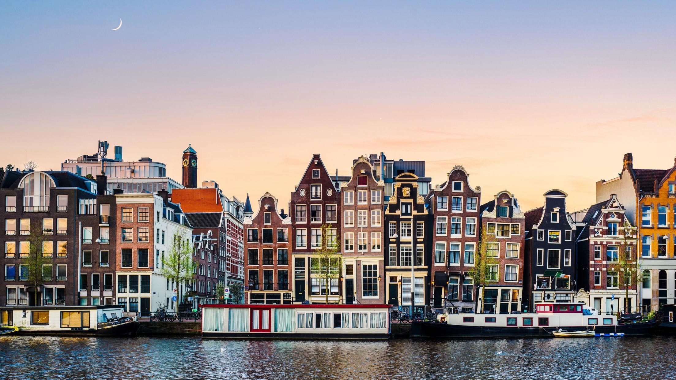 Uma fileira de casas coloridas &agrave; beira do canal em Amsterd&atilde;. Eles s&atilde;o tortos e parecem estar encostados um no outro.