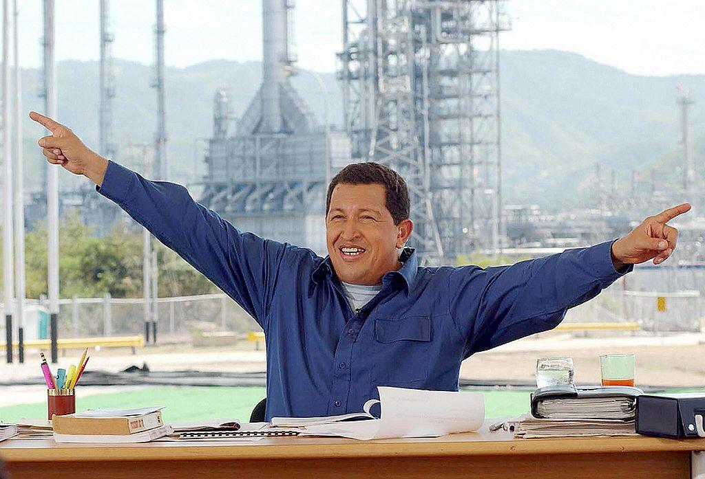 Hugo Chávez extiende los brazos sonriente con una planta industrial al fondo. 