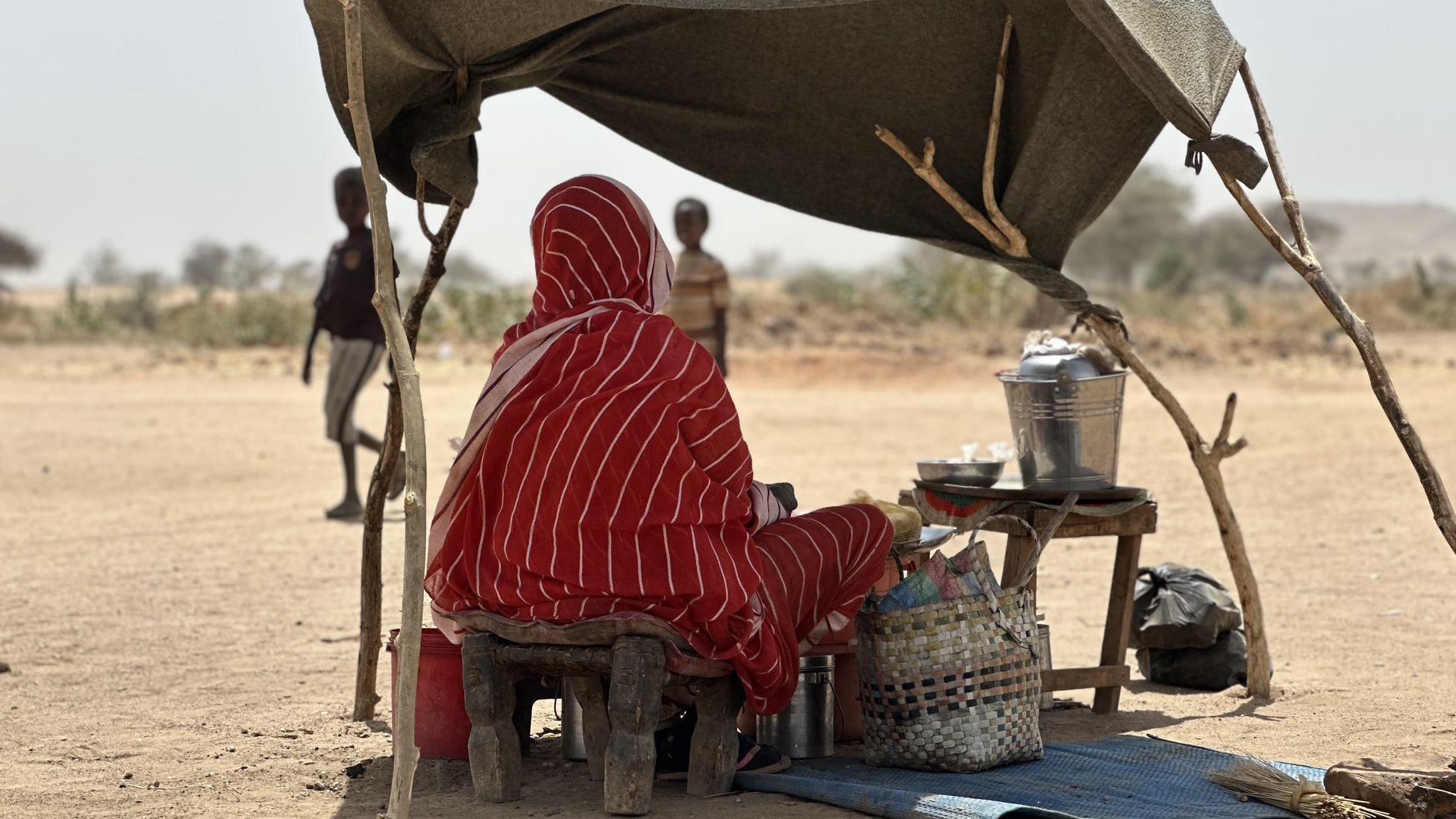 امرأة سودانية ترتدي ثوباً أحمر تجلس تحت مظلة بسيطة مصنوعة من القماش والأخشاب، وأمامها يظهر طفلان.