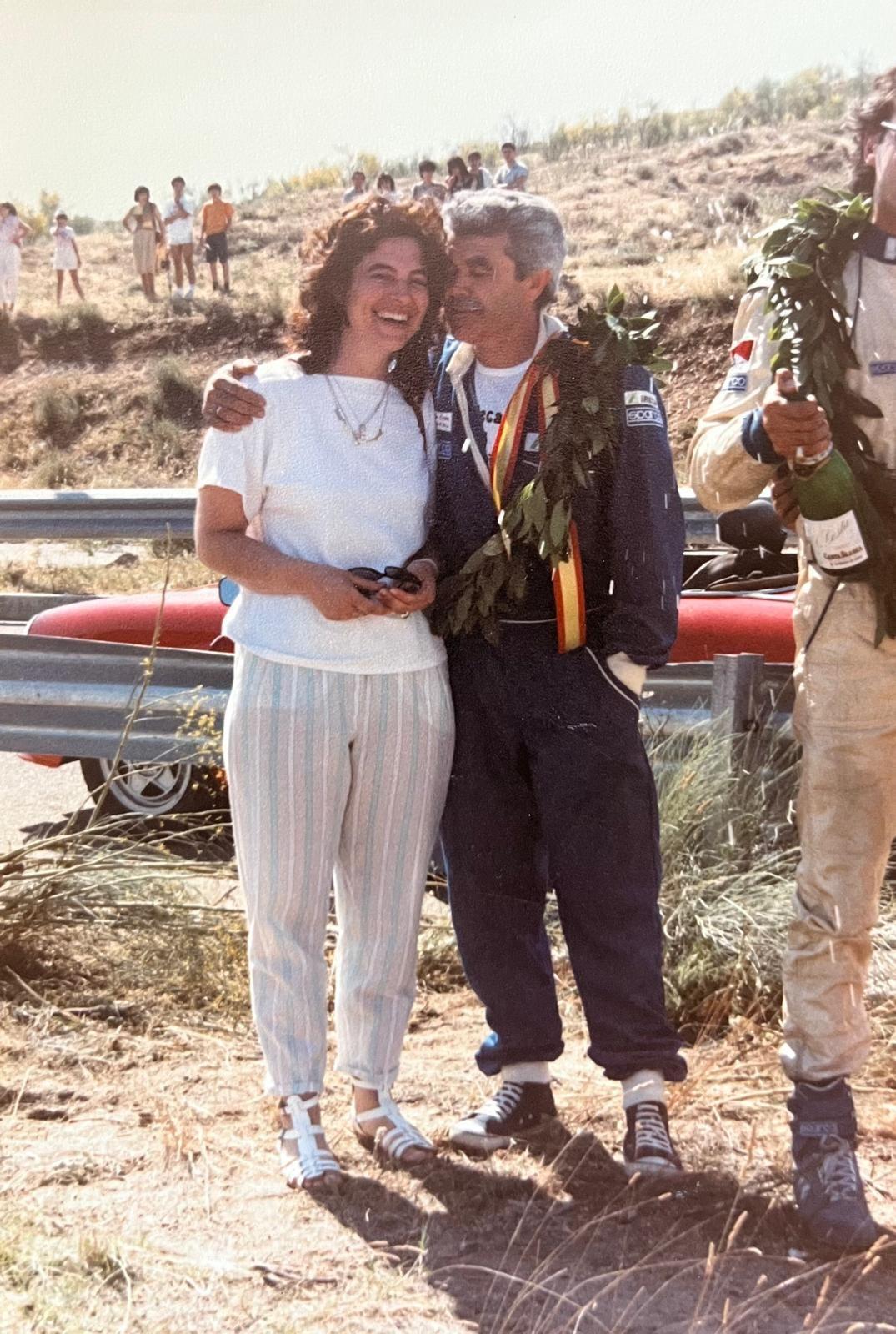 Juan, victorioso con una medalla al cuello y una corona de laureles, abraza con cariño a su esposa quien sonríe alegremente a la cámara.