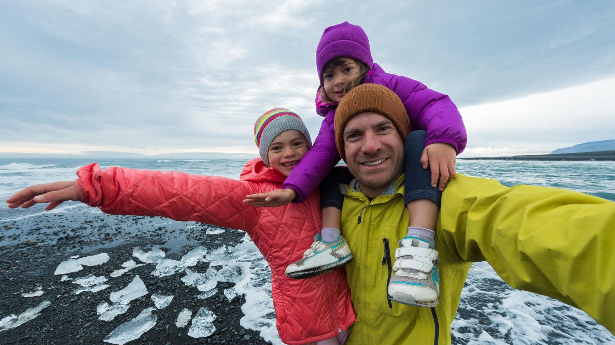 Un padre carga en sus hombros a una pequeña hija, mientras otra lo abraza, frente a un mar con hielo y un cielo invernal. Todos ríen.