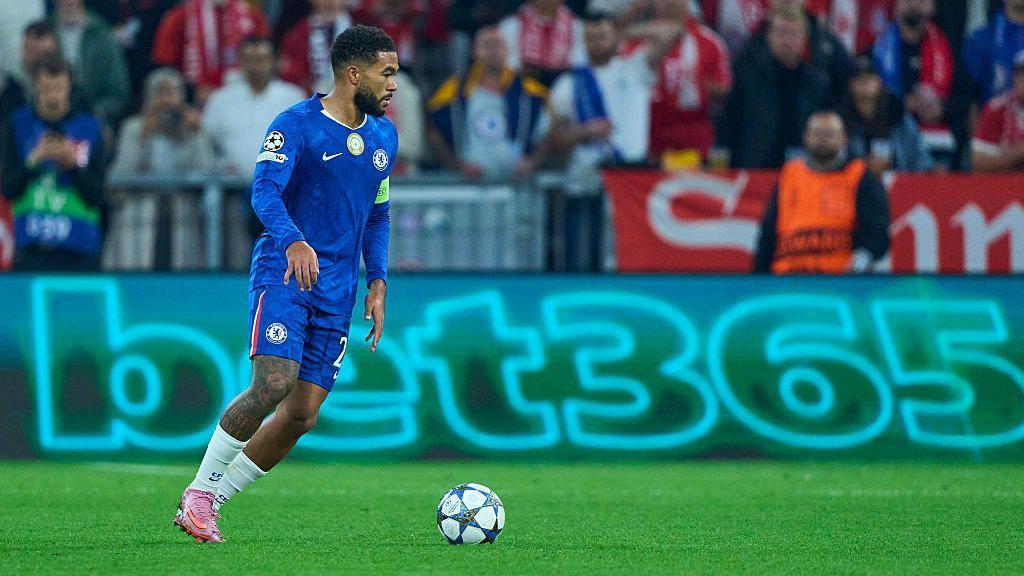 Jogador do Chelsea com a bola no gramado, com um an&uacute;ncio de bet no est&aacute;dio de futebol, ao fundo.