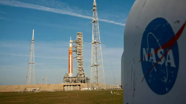 صاروخ مهمة أرتميس 2 البرتقالي يقف على منصة الإطلاق في قاعدة كينيد الفضائية وبجواره شعار وكالة ناسا قبل انطلاقه نحو القمر