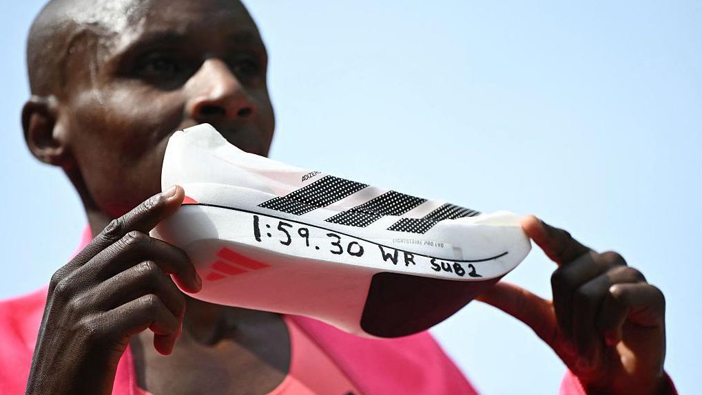 O queniano Sabastian Sawe posa com seu novo tempo de recorde mundial escrito em seu t&ecirc;nis de corrida na linha de chegada da Maratona de Londres de 2026