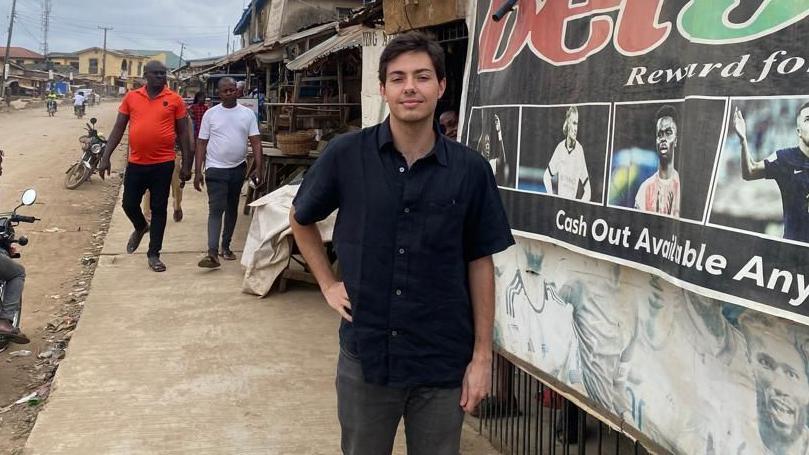 Carlos, un joven español de cabello oscuro, está de pie junto a un edificio al borde de la carretera con una gran pancarta publicitaria de apuestas deportivas. En la pancarta se aprecian claramente las palabras "bet9" y "Retiro de efectivo disponible en cualquier momento", junto a imágenes de futbolistas. Viste una camisa oscura de manga corta y pantalones oscuros. Se encuentra en un entorno urbano nigeriano, junto a una calle sin pavimentar, con peatones y un motociclista detrás de él, y dos grandes antenas parabólicas instaladas sobre el cartel.