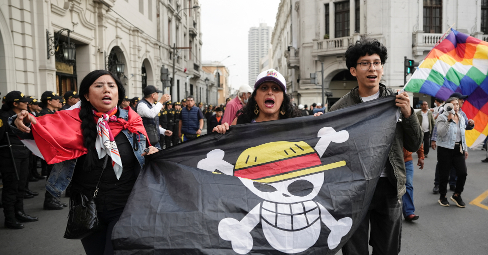 Tres manifestantes, dos mujeres y un hombre, sujetan una bandera negra con una calavera con dos tibias y un sombrero de la serie de animación "One piece" en la plaza de San Martín, en Lima