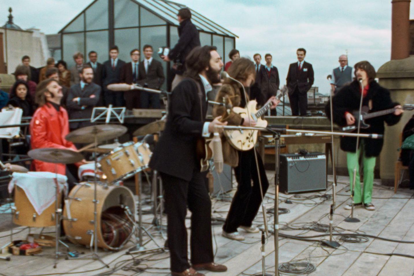Os Beatles tocam no telhado do n&uacute;mero 3 da Savile Row. Paul McCartney est&aacute; em primeiro plano, debru&ccedil;ado sobre o microfone, enquanto Ringo toca bateria vestindo uma jaqueta de couro vermelha. Atr&aacute;s deles est&atilde;o John Lennon e George Harrison, sob o olhar de dezenas de homens de terno.