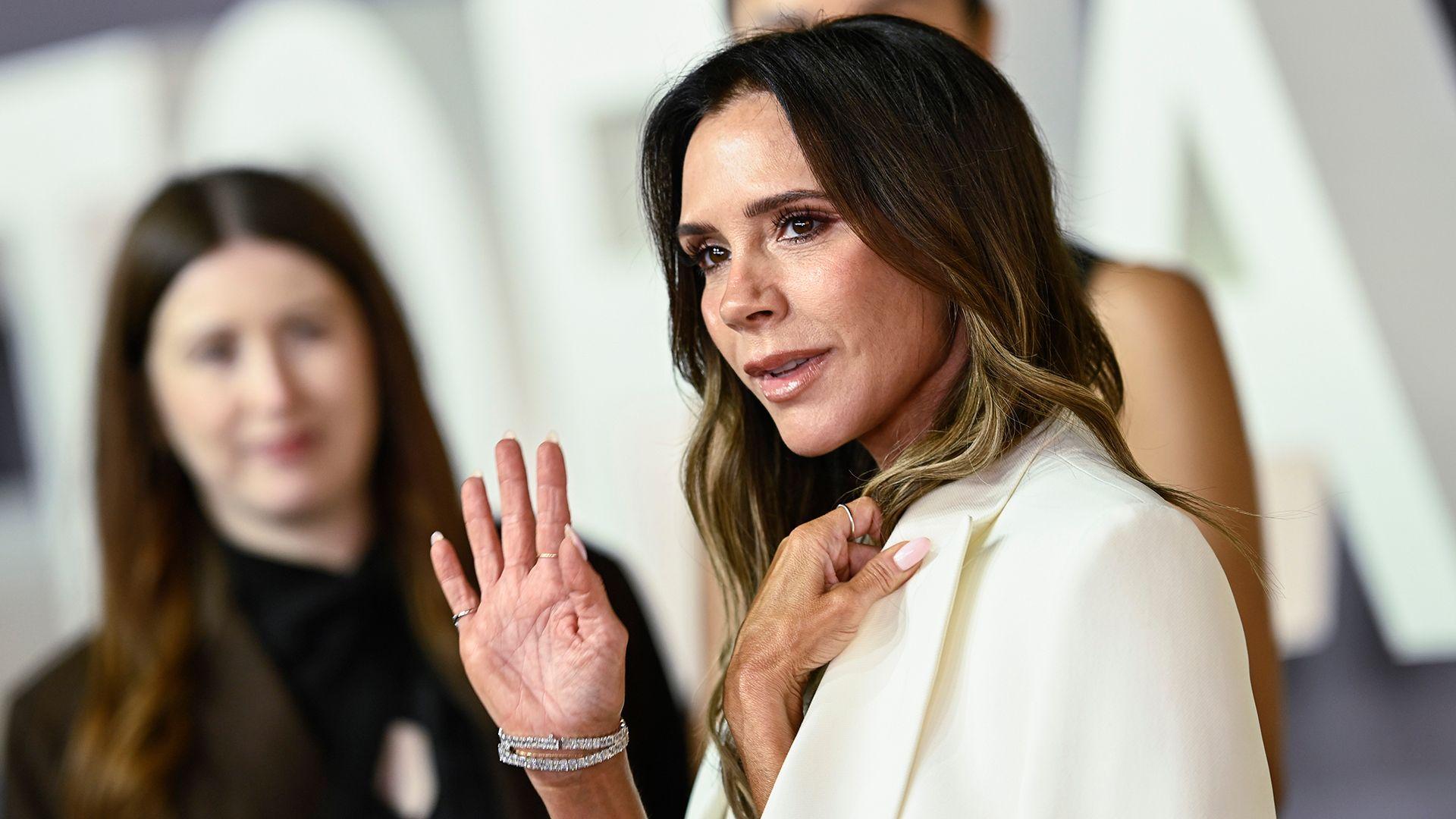 Victoria Beckham saluda; viste un traje blanco y hay otras personas de fondo.