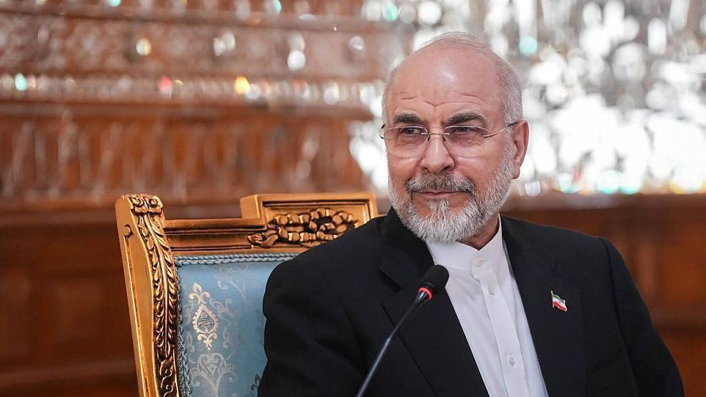رئيس البرلمان الإيراني محمد باقر قاليباف، مرتدياً بدلة داكنة وقميصاً أبيض وشارة على شكل العلم الإيراني على ياقة سترته، يجلس على كرسي مزخرف أمام ميكروفون.
