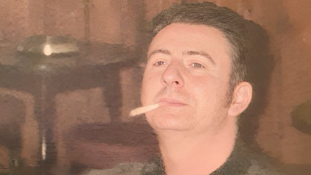 Danny est&aacute; sentado em um ambiente interno que parece ser um pub, usando um su&eacute;ter escuro estampado. Ele tem cabelo curto e escuro e olha levemente para cima, com um cigarro na boca