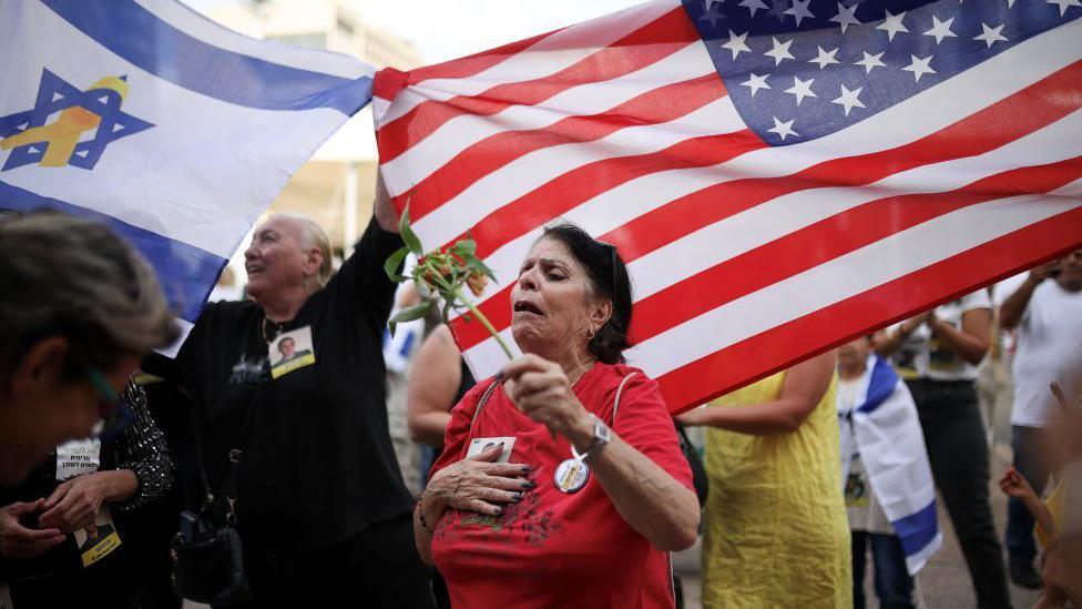 Žena u crvenoj majici drži cvet preplavljena emocijama. Iza nje su ljudi koji mašu izraelskim i američkim zastavama.