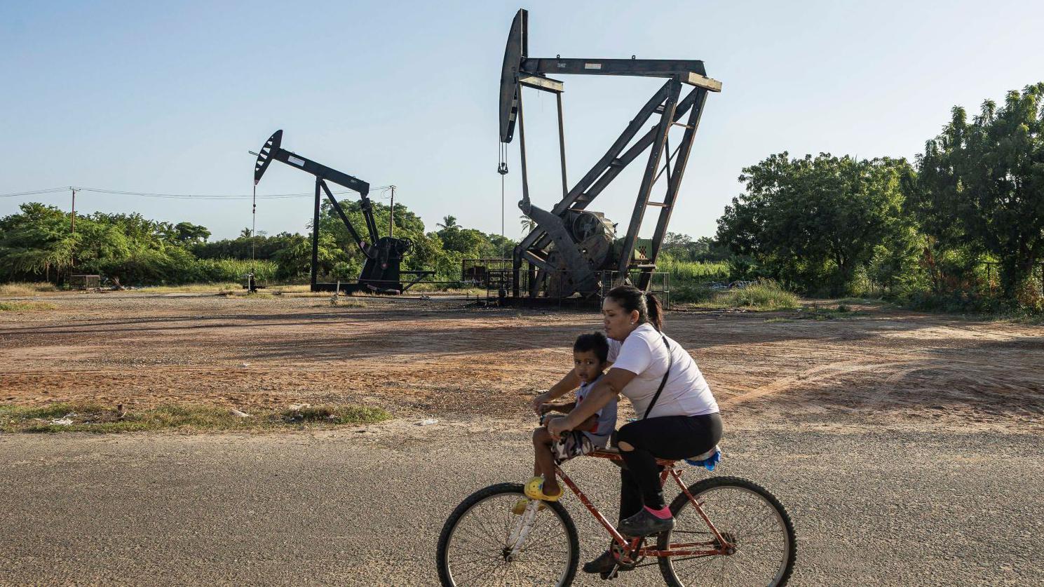 A woman rides a bicycle with a child in front of oil derricks in Cabimas, Venezuela, 18 December 2025. US President Donald Trump announced a total blockade of all sanctioned oil tankers entering and leaving the country, a strategy analysts say is intended to be selective and part of growing pressure over crude oil and other natural resources.