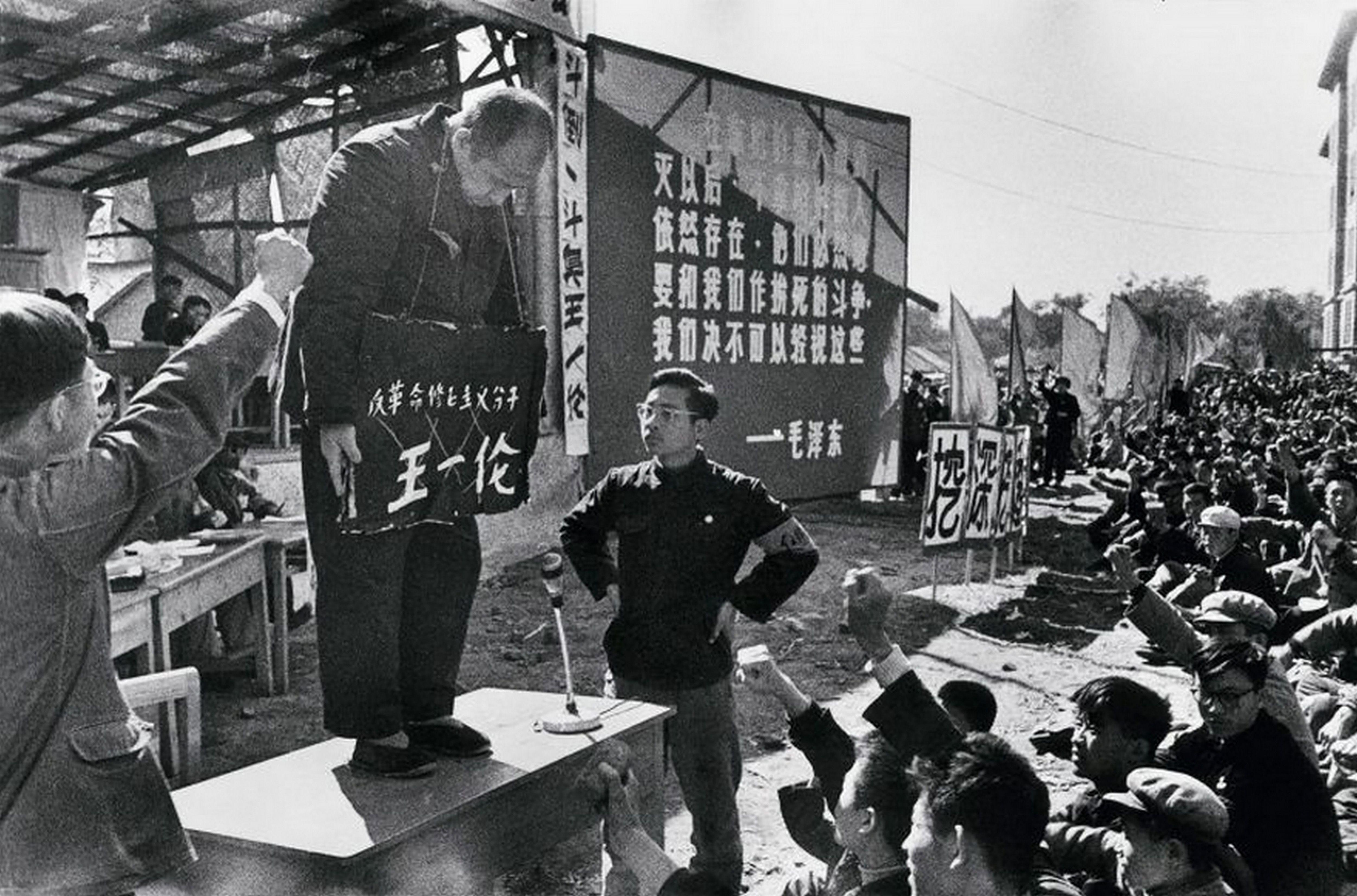 文革批鬥現場，一名長者站在桌上，頸掛一個寫有其姓名的牌子，周圍有群眾握拳。