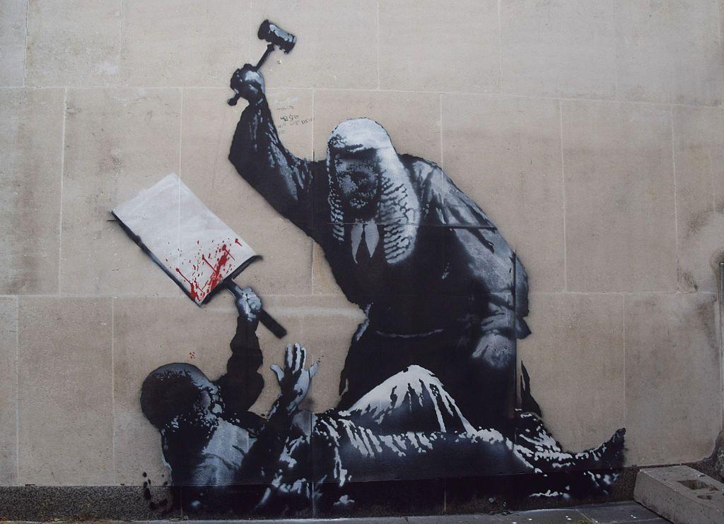 La última obra del artista callejero Banksy aparece en un muro de los Reales Tribunales de Justicia, representando a un juez golpeando a un manifestante con un mazo.