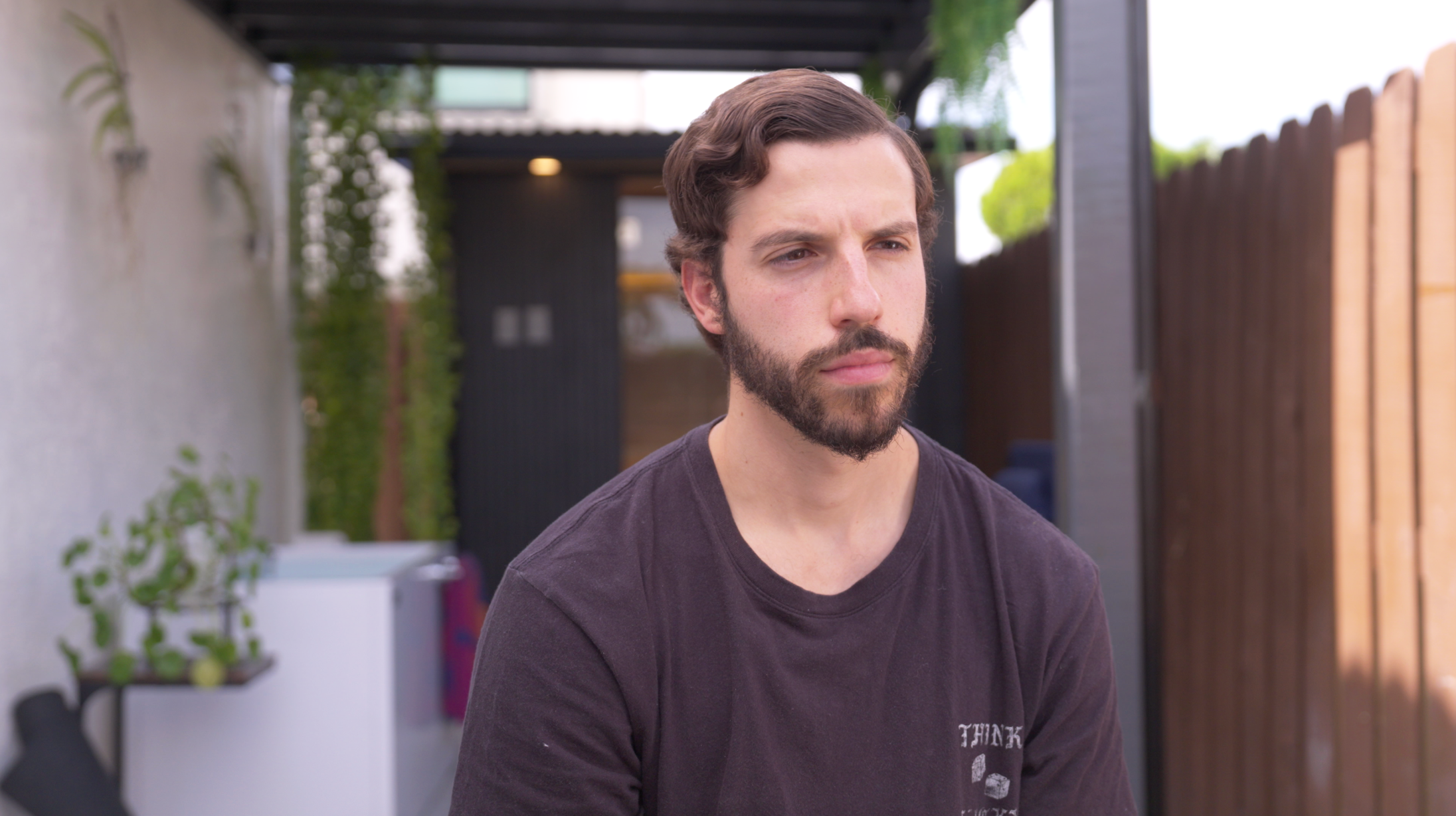 Simon sits in his back garden, looking to the side of the camera. He has a dark beard and dark short wavy hair. He is wearing a dark grey t-shirt. 
