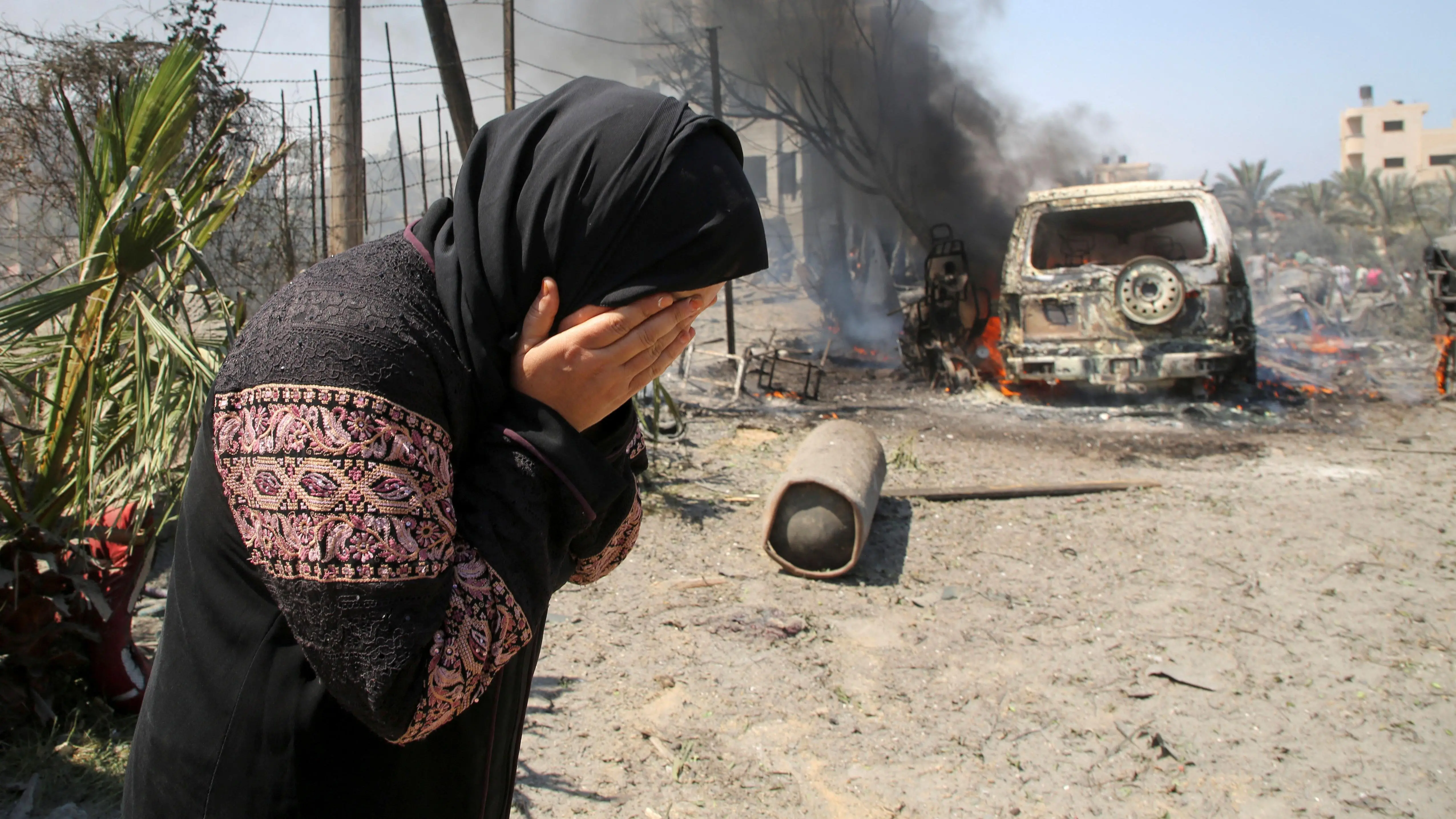 سيدة تغطي وجهها بكفيها وتظهر خلفها مركبة محترقة في منطقة مواصي غزة