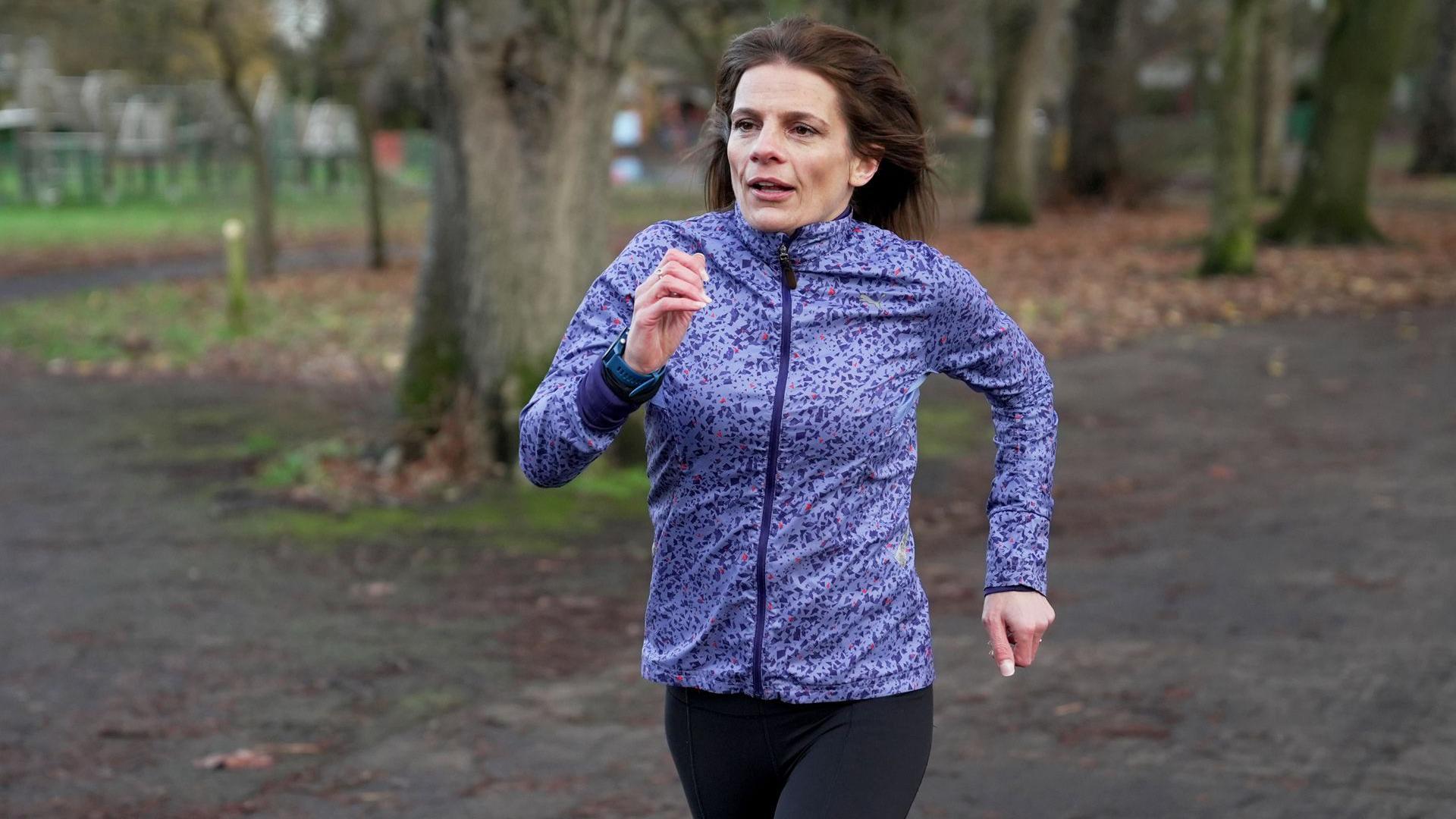 Ruth Clegg, uma mulher de cabelos castanhos e jaqueta esportiva roxa, corre em uma pista que atravessa um parque.