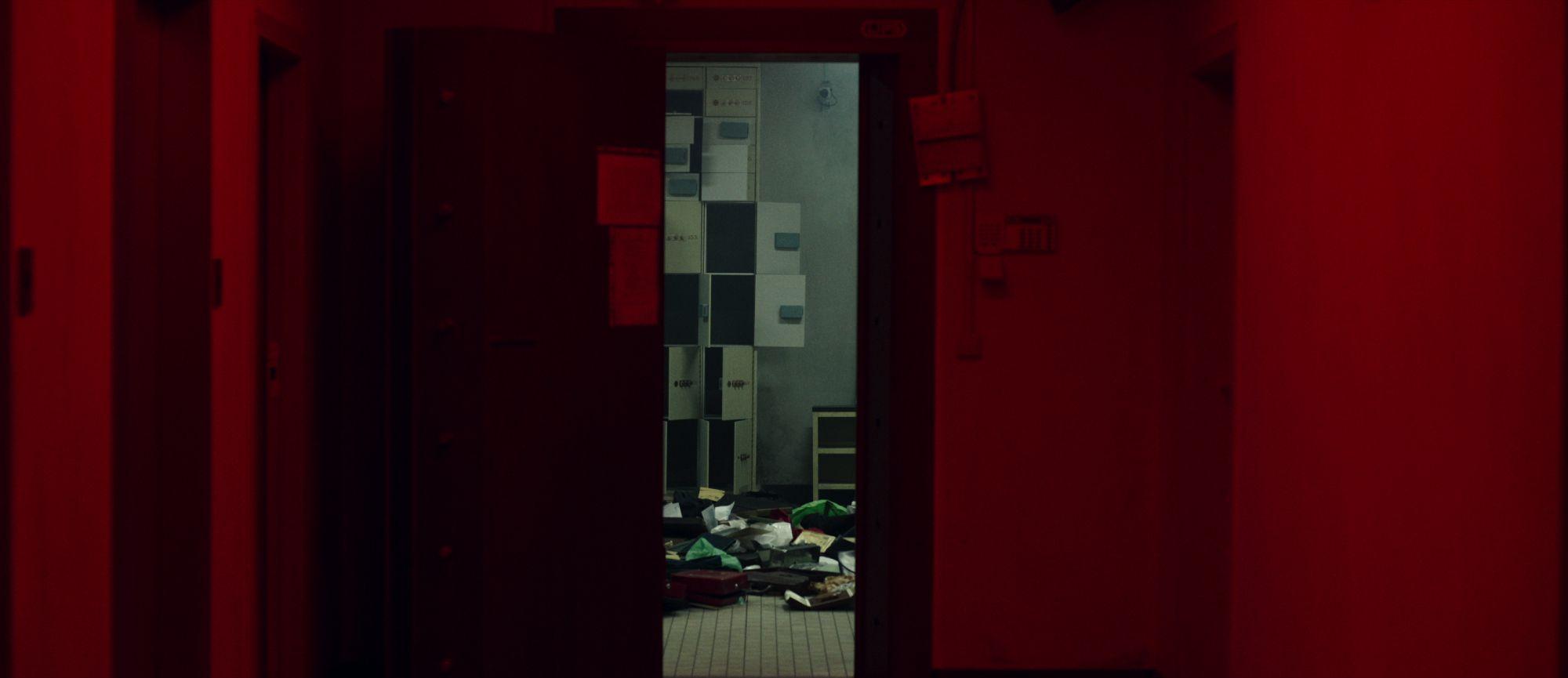 Pasillo iluminado en rojo con una puerta abierta que muestra una sala con casilleros y papeles esparcidos en el suelo