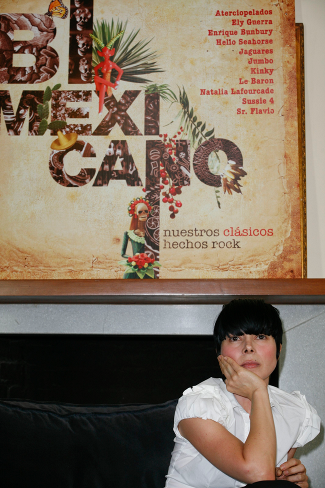 Ely Guerra, con una camisa blanca, está sentada con la mano en la barbilla. Detrás tiene la portada del álbum que dice “Bimexicano, Nuestros Clásicos Hechos Rock”.