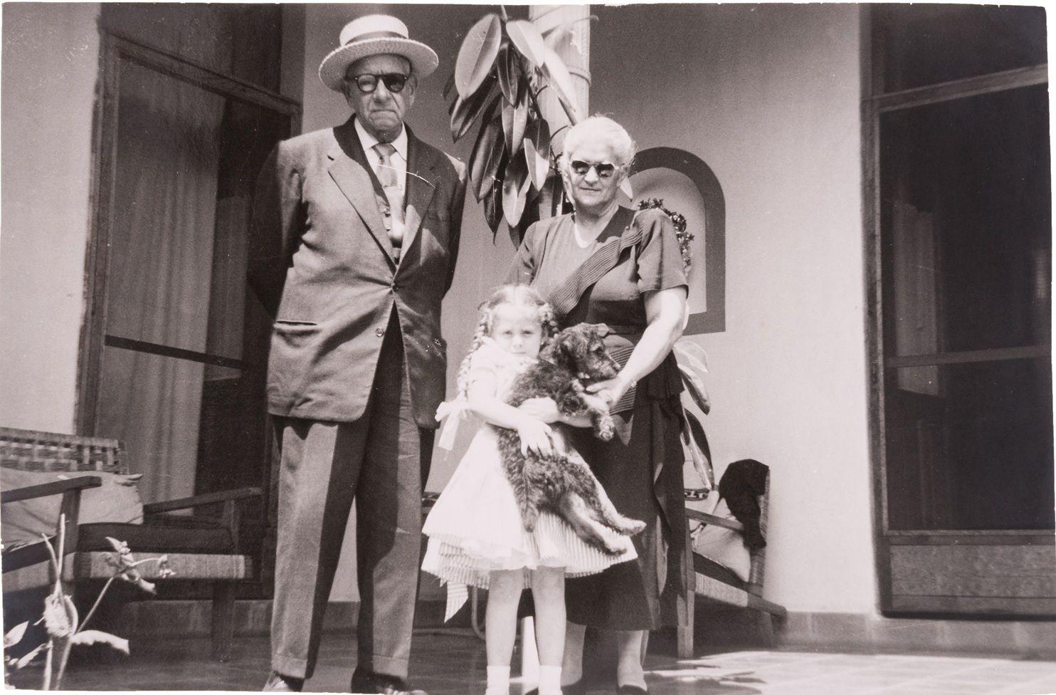 Alberto Veraza, vestido de traje, con corbata y sombrero, tiene las manos hacia atrás. A su lado está Adriana, quien tiene el cabello blanco, lleva lentes y un vestido oscuro. Muy cerca de ella está Mara, que tiene trenzas y un vestido blanco y carga un perro. Los tres miran a la cámara y están en lo que parece ser el patio de una casa
