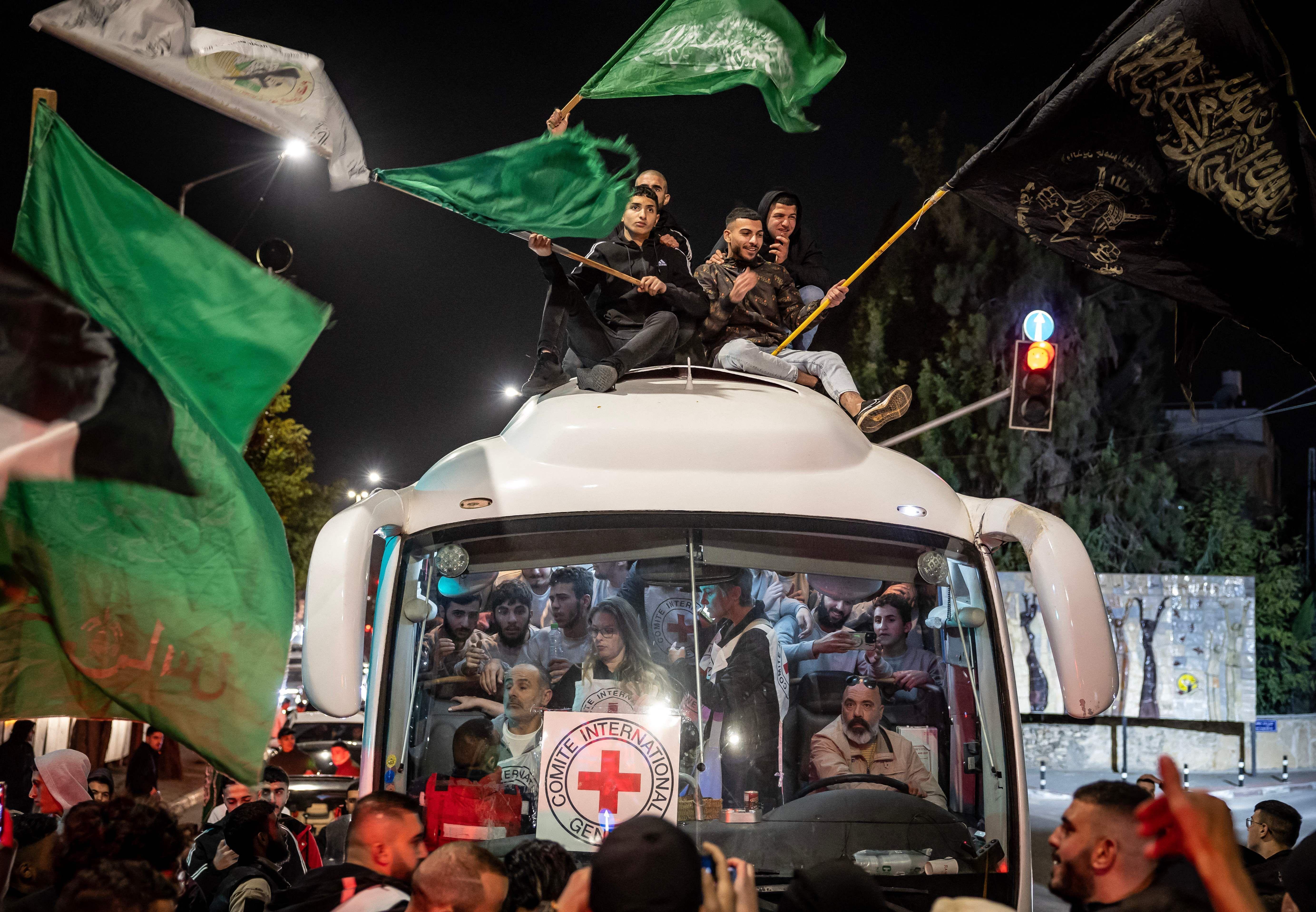 حافلة بيضاء مليئة بفلسطينيين مفرج عنهم من السجون الإسرائيلية، على زجاجها الأمامي إشارة الصليب الأحمر، البعض يجلس على ظهر الحافلة ويحمل عدد منهم العلم الأخضر لحركة حماس في أجواء احتفالية.