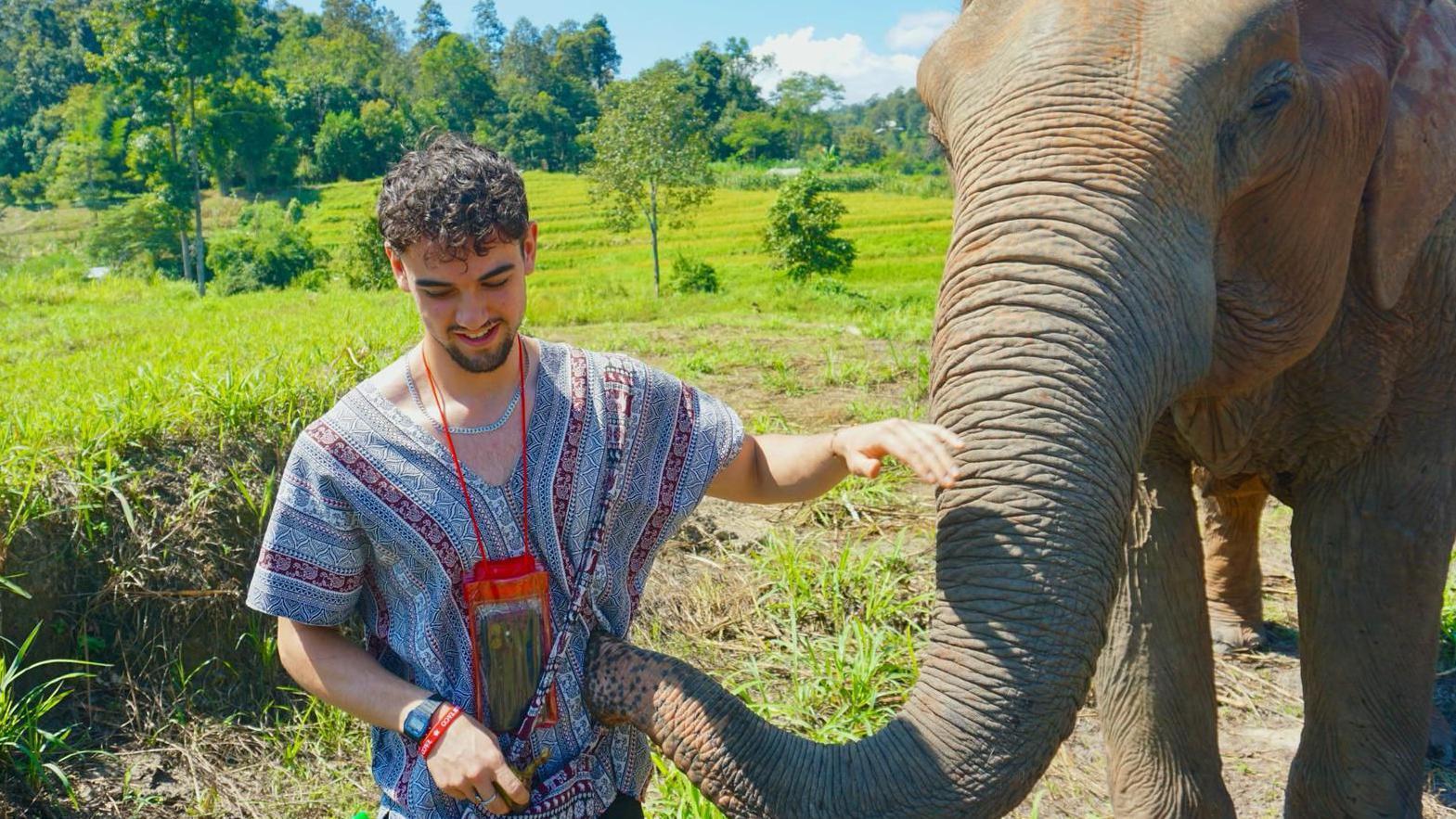 Calum Macdonald acaricia a un elefante mientras este le toca la camisa a rayas con la trompa. Detrás de ellos se ven arrozales verdes y, más allá, bosques tropicales. Tiene el pelo corto y rizado de color negro.