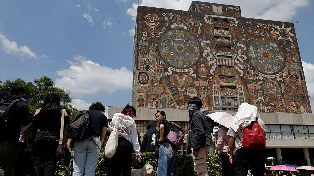 Un grupo de estudiantes frente al edificio principal de la universidad autónoma de méxico. El edificio tiene murales con elementos de la cultura mexicana. 