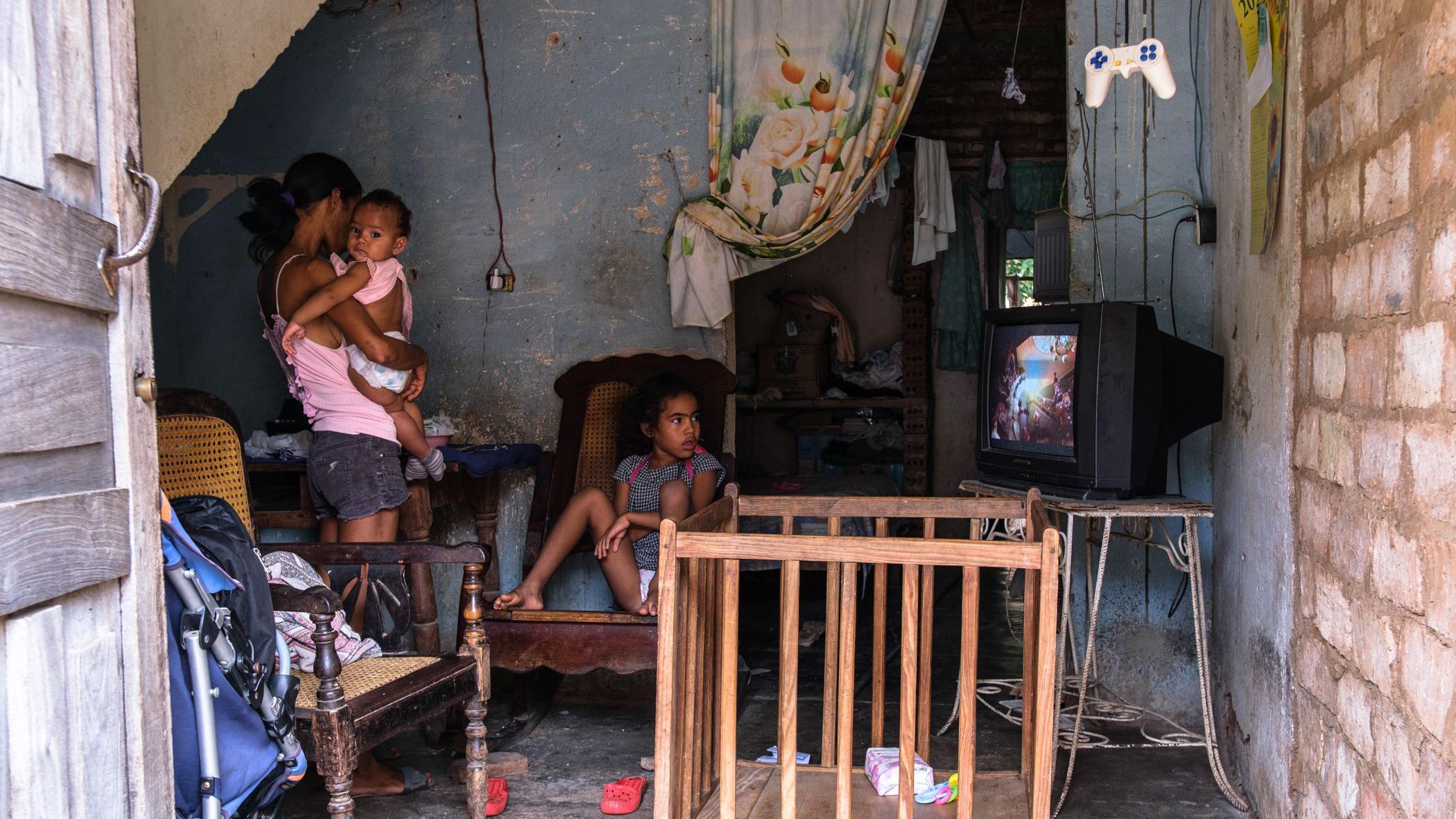Mulher com duas crianças em uma casa em Trinidad, Cuba. Elas estão assistindo televisão. Há duas cadeiras e um berço