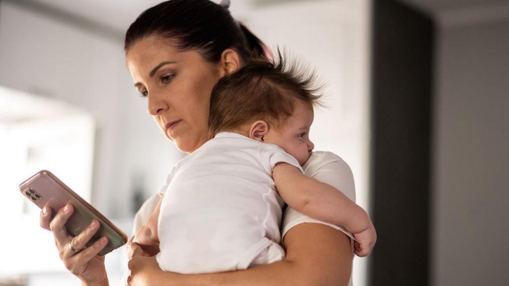 žena sa bebom u naručju drži u ruci mobilni telefon